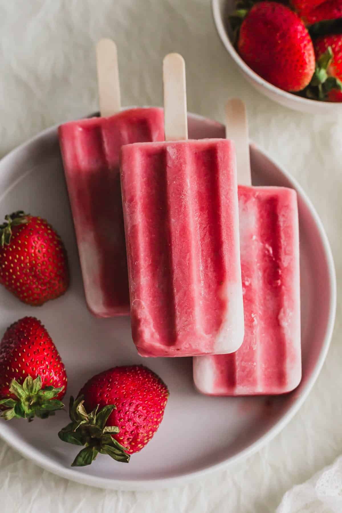 3 Strawberry sago popsicles piled on a white plate, with strawberries on the side.